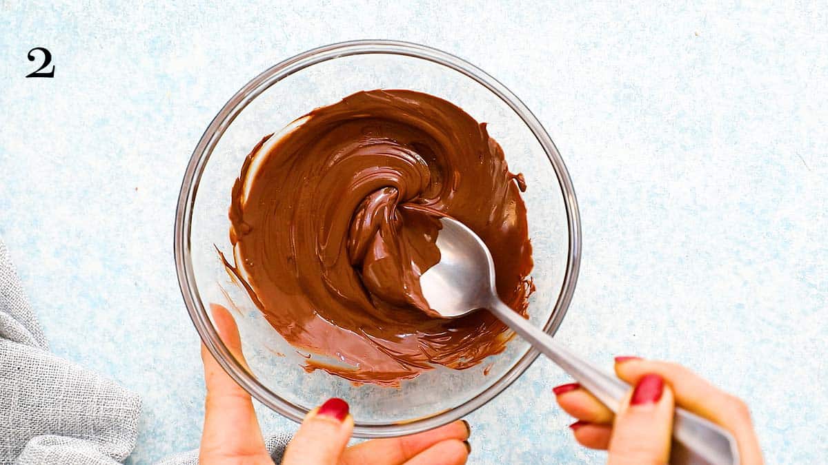 two hands stirring melted chocolate in a glass bowl using a spoon.