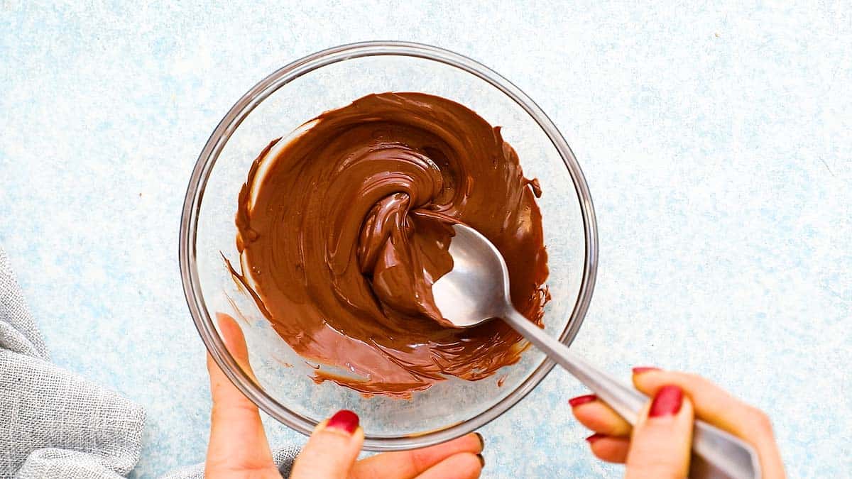 2 hands mixing melted chocolate along with a spoon in a bowl. 