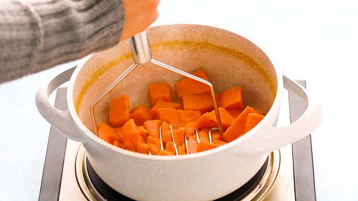 a hand using a potato masher to mash sweet potatoes.