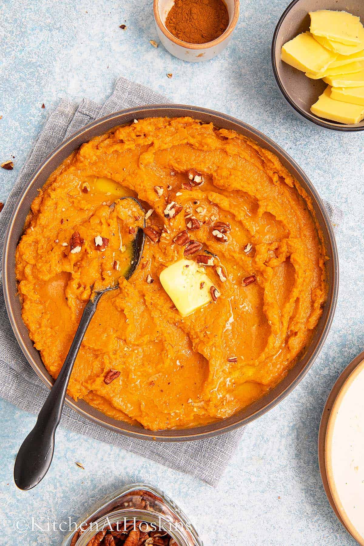  round platter filled with mashed sweet potatoes topped with butter, pecans along with a spoon.