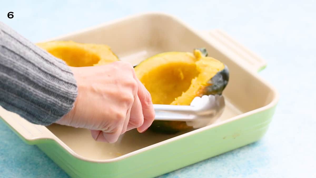 a hand flipping a cooked acorn squash halve with a tongs.