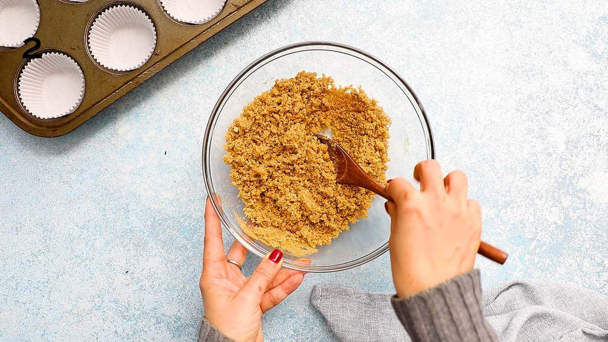 two hands mixing graham cracker crumbs in a glass bowl with wooden spoon.