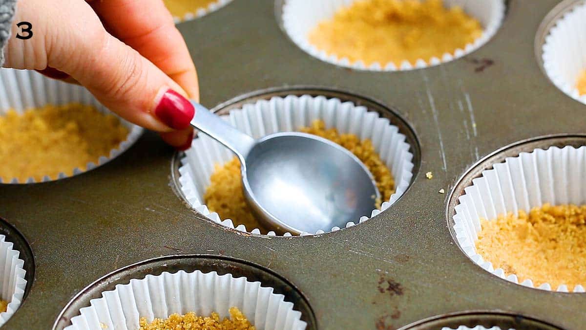 a hand pressing cracker crumbs into a paper lined muffin pan using a stain less steel tablespoon.