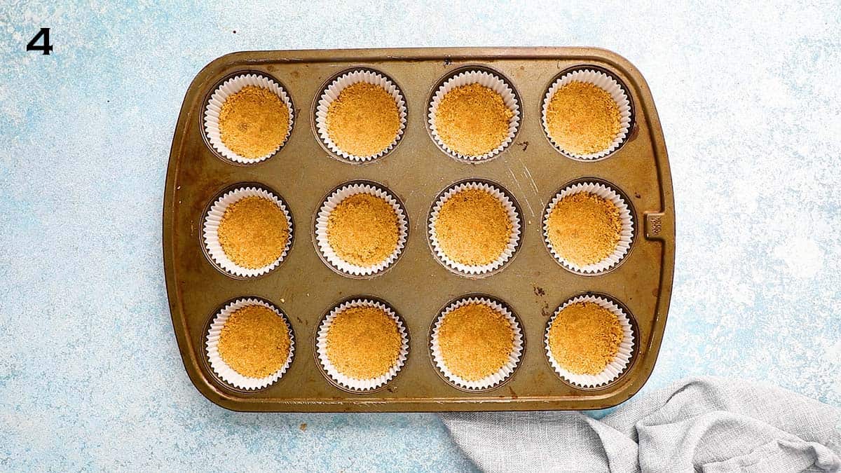 a 12-cup muffin pan, lined with white paper lined, which are filled with graham cracker crumbs.