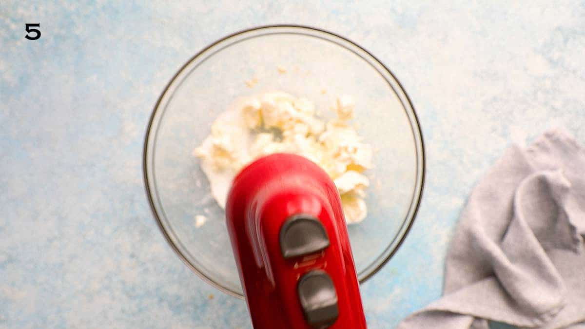 red electric beater creaming cream cheese in a glass bowl.