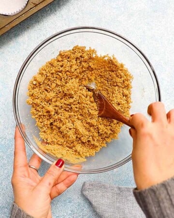 two hands mixing brown graham cracker crust in a glass bowl.