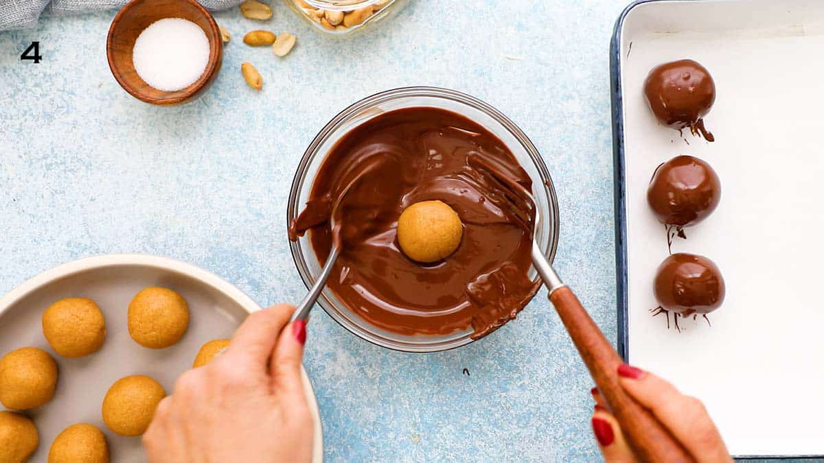 two hands dipping a peanut butter ball in a bowl of melted chocolate, using a fork and spoon.