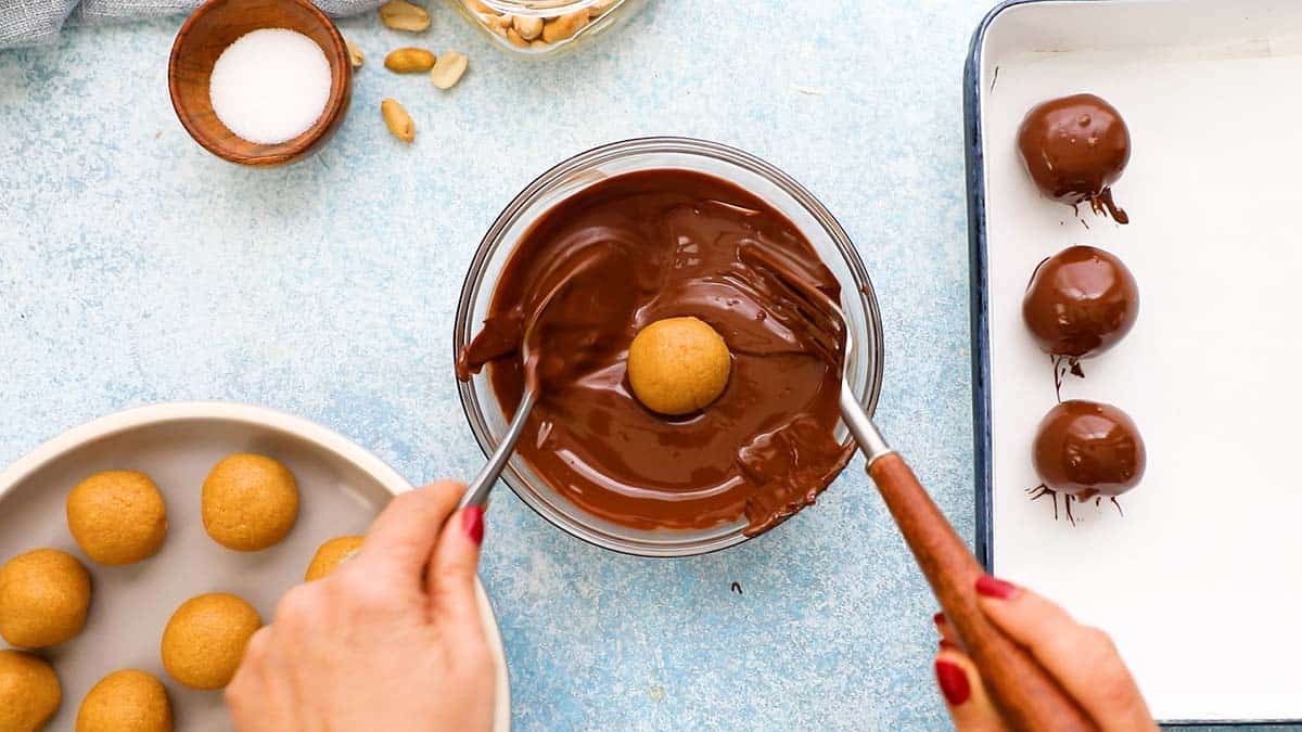 two hands dipping peanut butter ball in a bowl of melted chocolate.