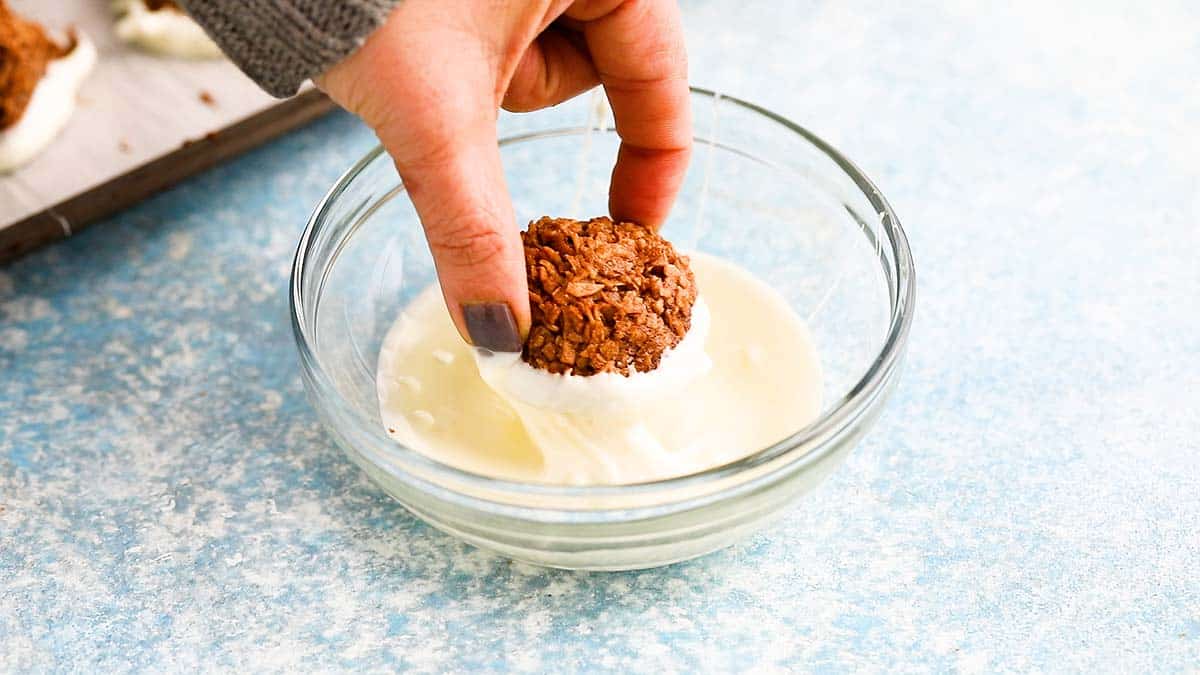 a hand dipping a chocolate macaroon in melted white chocolate.
