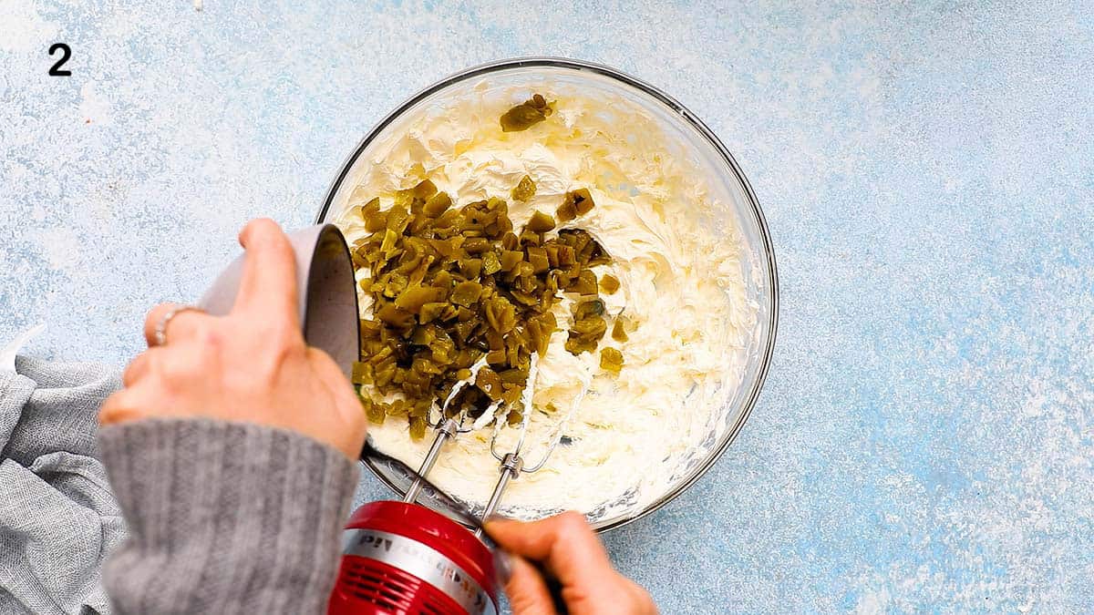two hands adding canned jalapenos into a bowl of cream cheese.