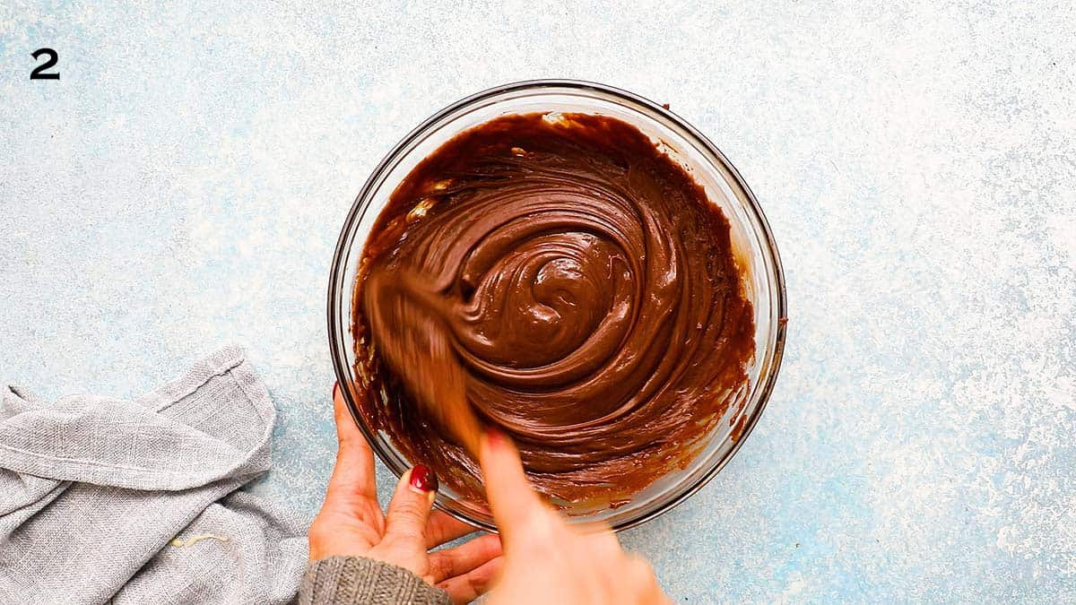 a hand stirring melted chocolate fudge mixture in a glass bowl using a wooden spoon.