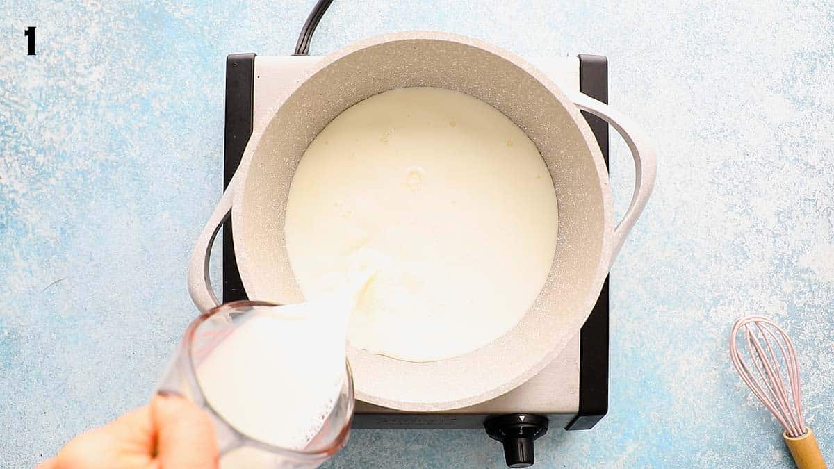 a hand pouring milk into a white saucepan placed on a stovetop.