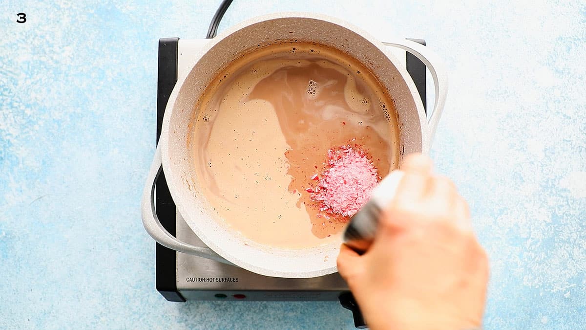a hand adding crushed candy canes into a white saucepan placed on a stovetop.