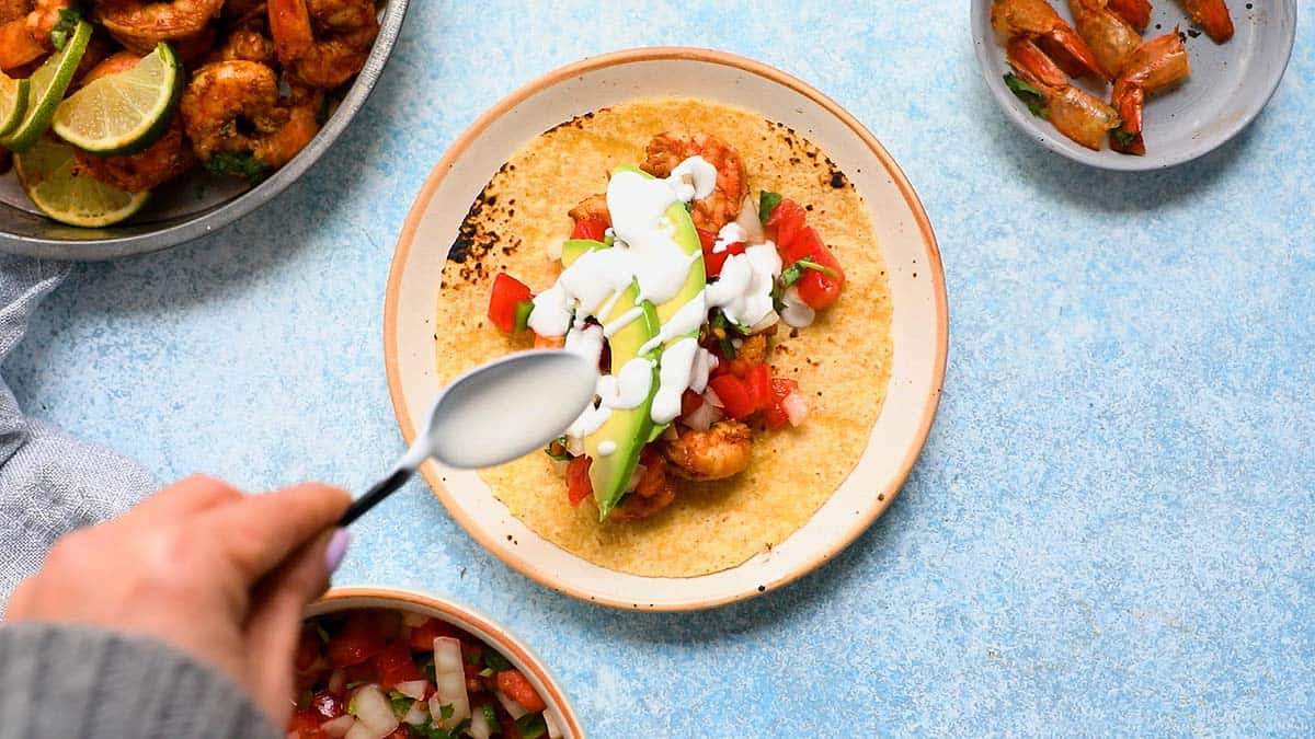 a hand drizzling sour cream sauce on a taco with avocado slices.