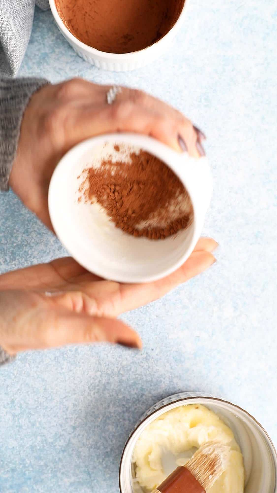 two hands dusting cocoa powder inside a ramekin.