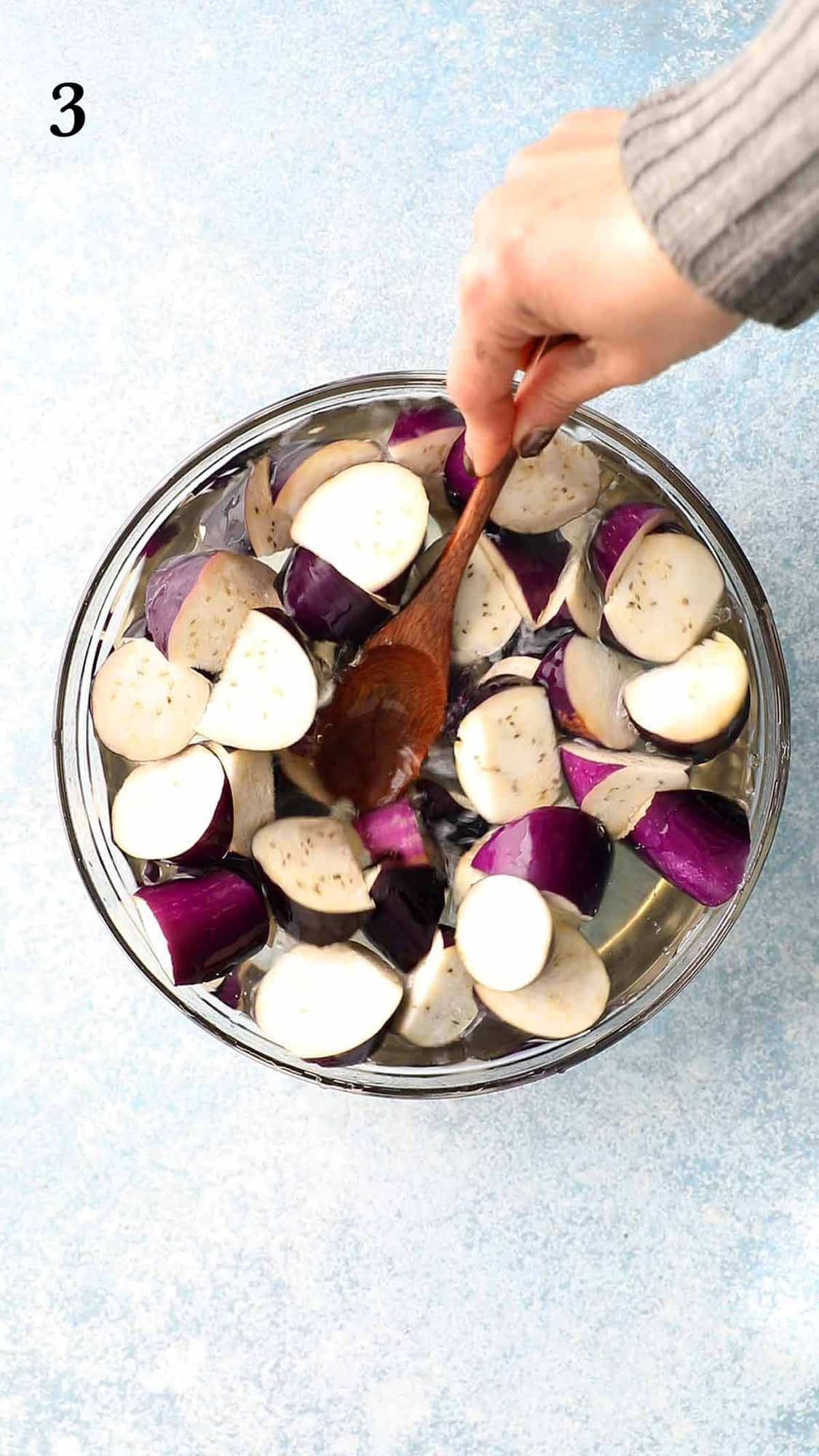 a hand stirring eggplant chunks in in a glass bowl.
