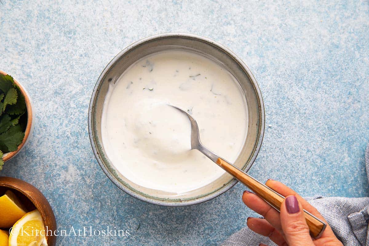a hand holding a metal spoon with creamy white sauce in a blue bowl.