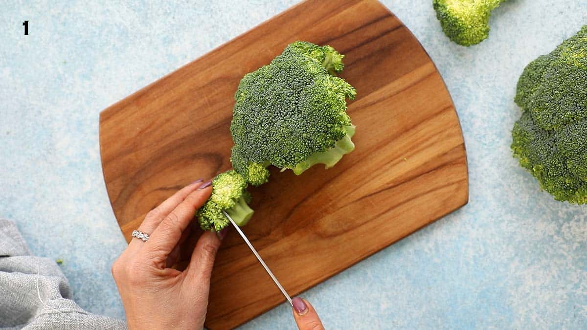 two hands chopping a head of broccoli on a wooden board using a knife.