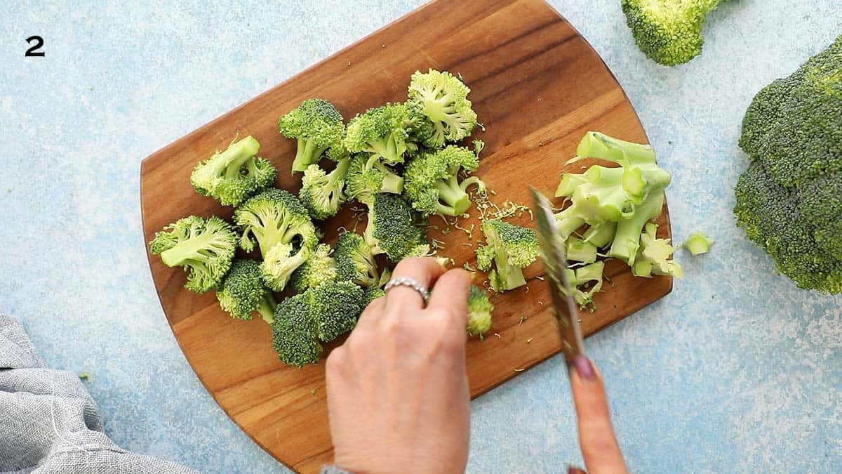 two hands chopping a broccoli into florets, on a wooden board using a knife.
