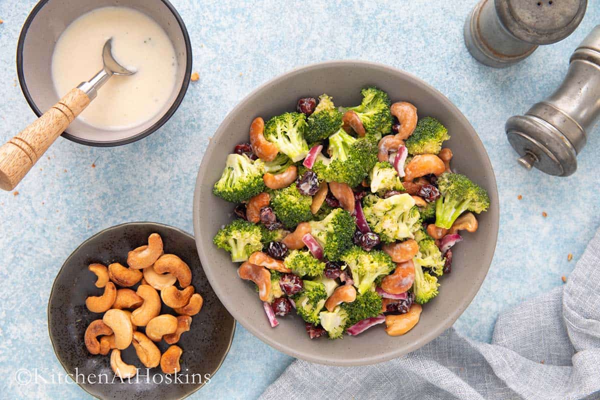 grey bowl with broccoli salad, small black plate with cashews, salt and pepper shakers on the side.