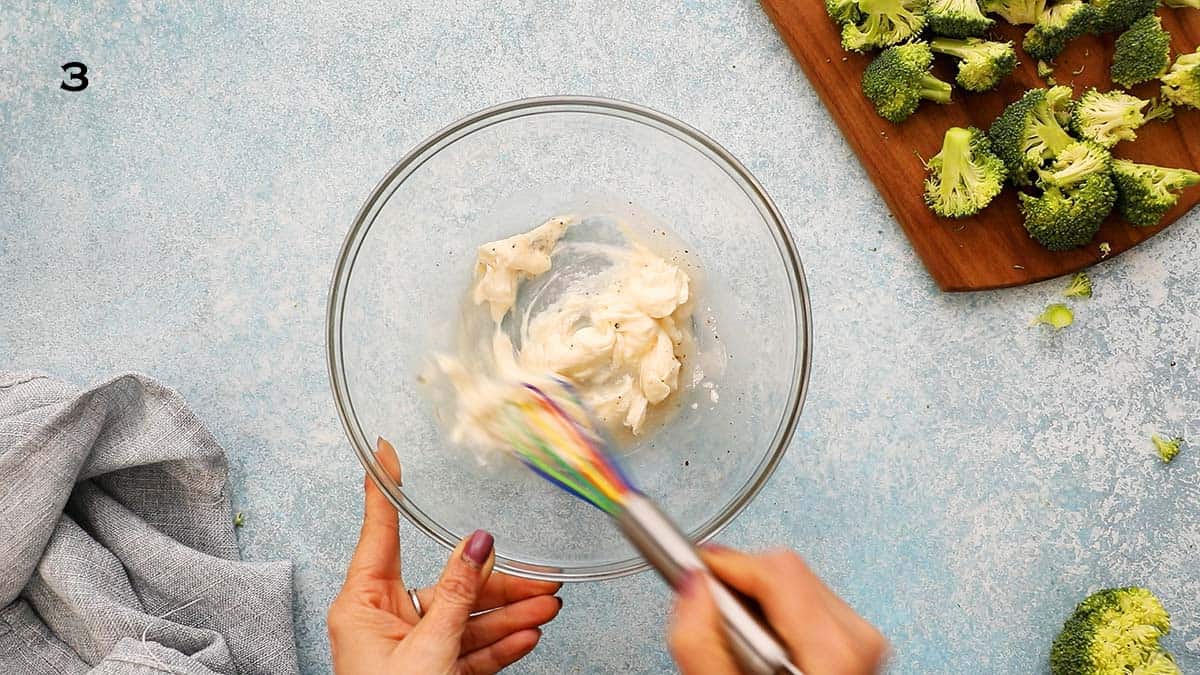 2 hands whisking white sauce in a glass bowl using a multi colored whisk.