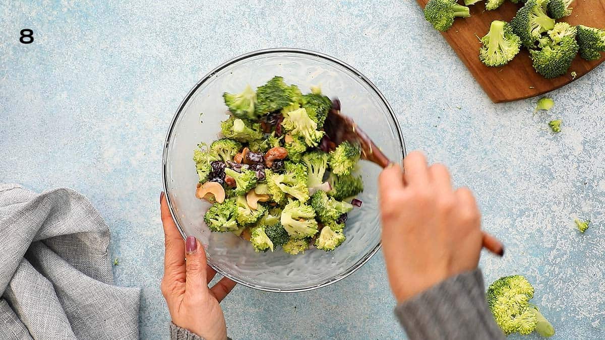 2 hands tossing broccoli salad in a glass bowl with a wooden spoon.