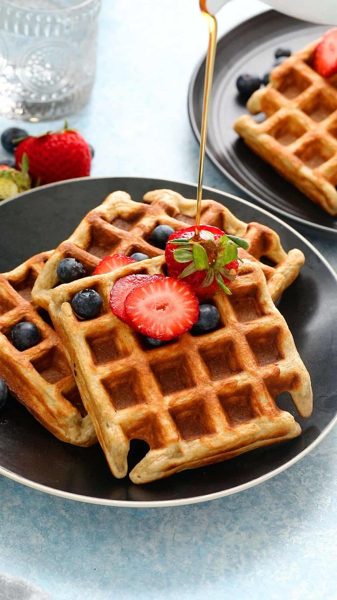 3 waffles on a black plate topped with berries and maple syrup.