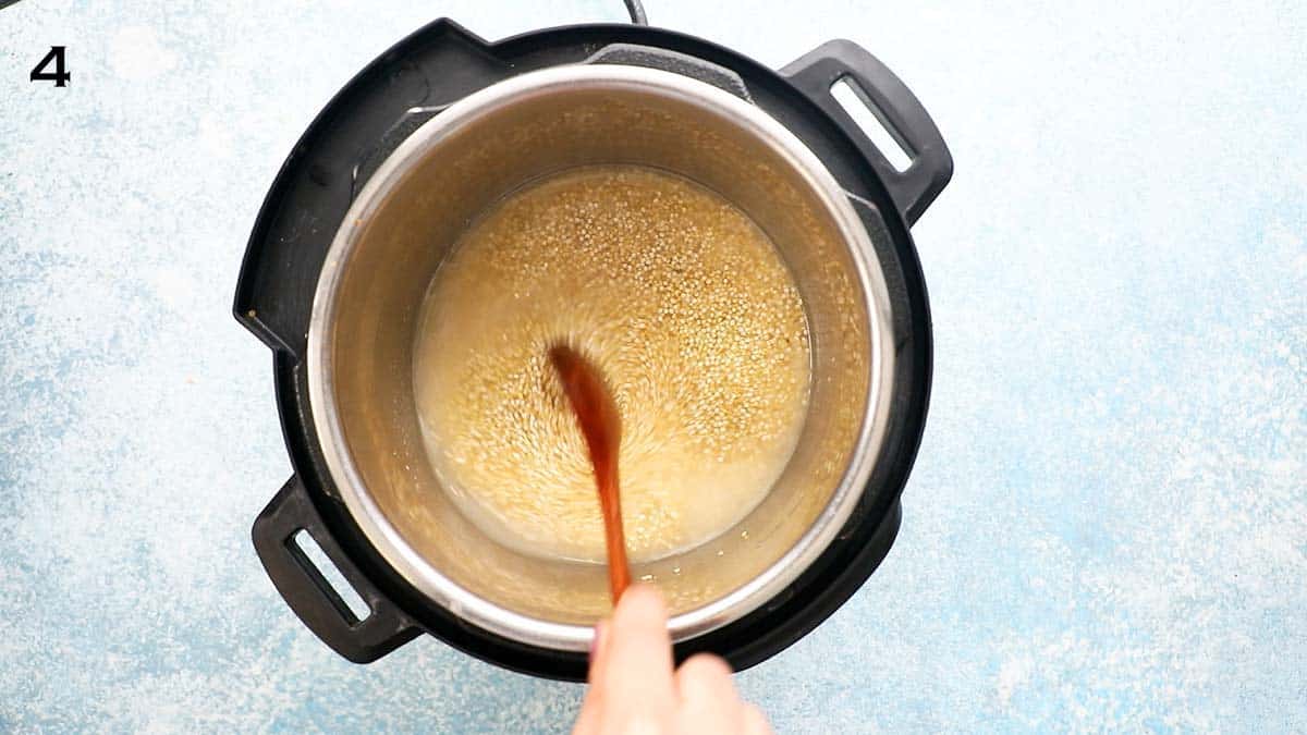 a hand stirring raw quinoa and water in an instant pot with a wooden spoon.