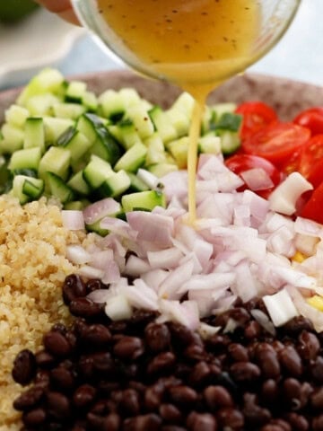 a hand pouring honey lime dressing over quinoa salad in a brown bowl.