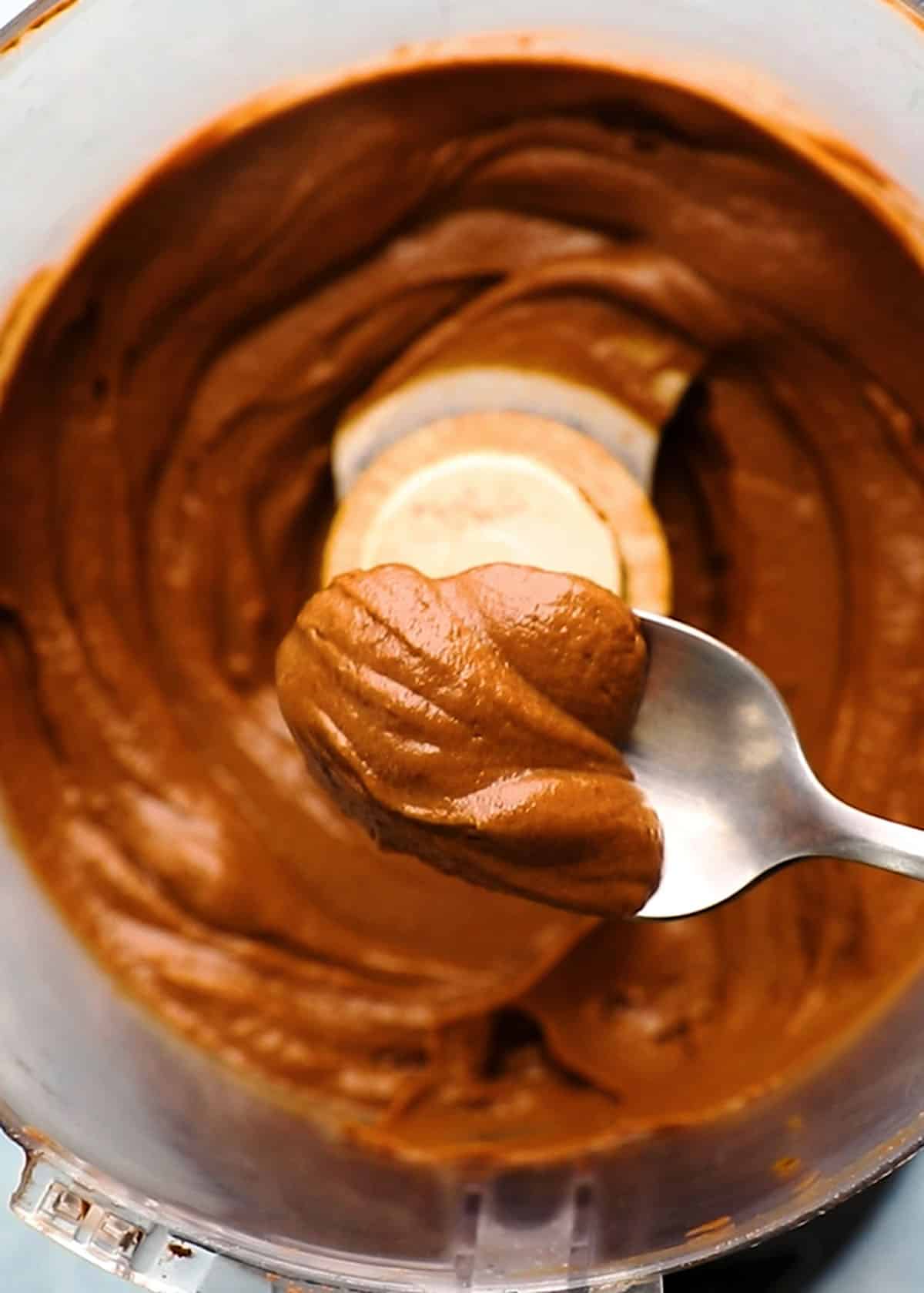 a hand holing a spoonful of brown chocolate avocado mousse above a food processor.