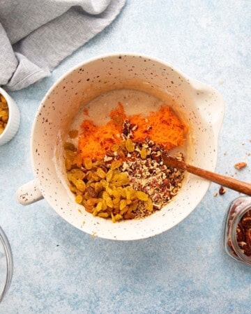 grated carrots, pecans and raisins in a white bowl along with a wooden spoon.