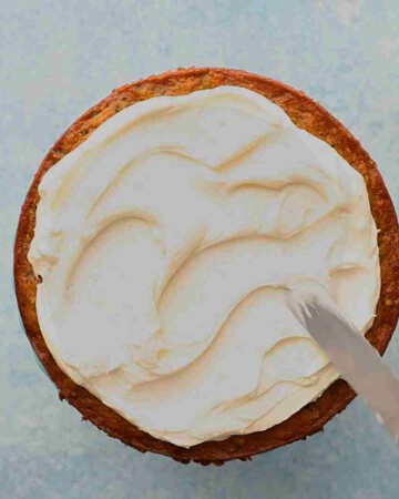 a hand spreading white frosting on a round carrot cake. 