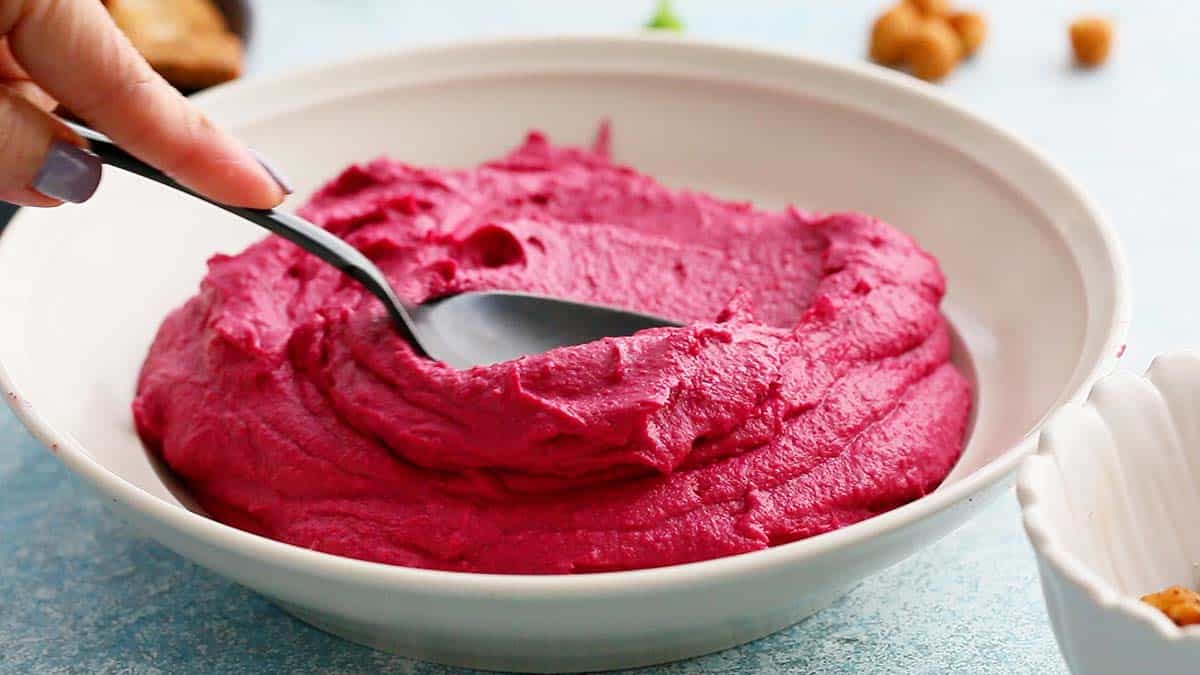 a black spoon swirling pink hummus in a white bowl.