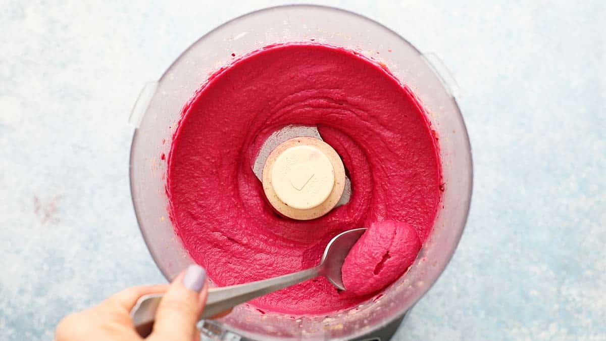 a spoon scooping out some pink hummus from a food processor.