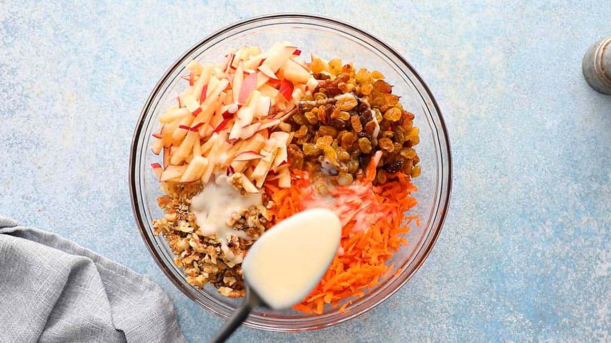 a black spoon drizzling creamy white dressing into a glass bowl with grated carrots, raisins and chopped apples. 
