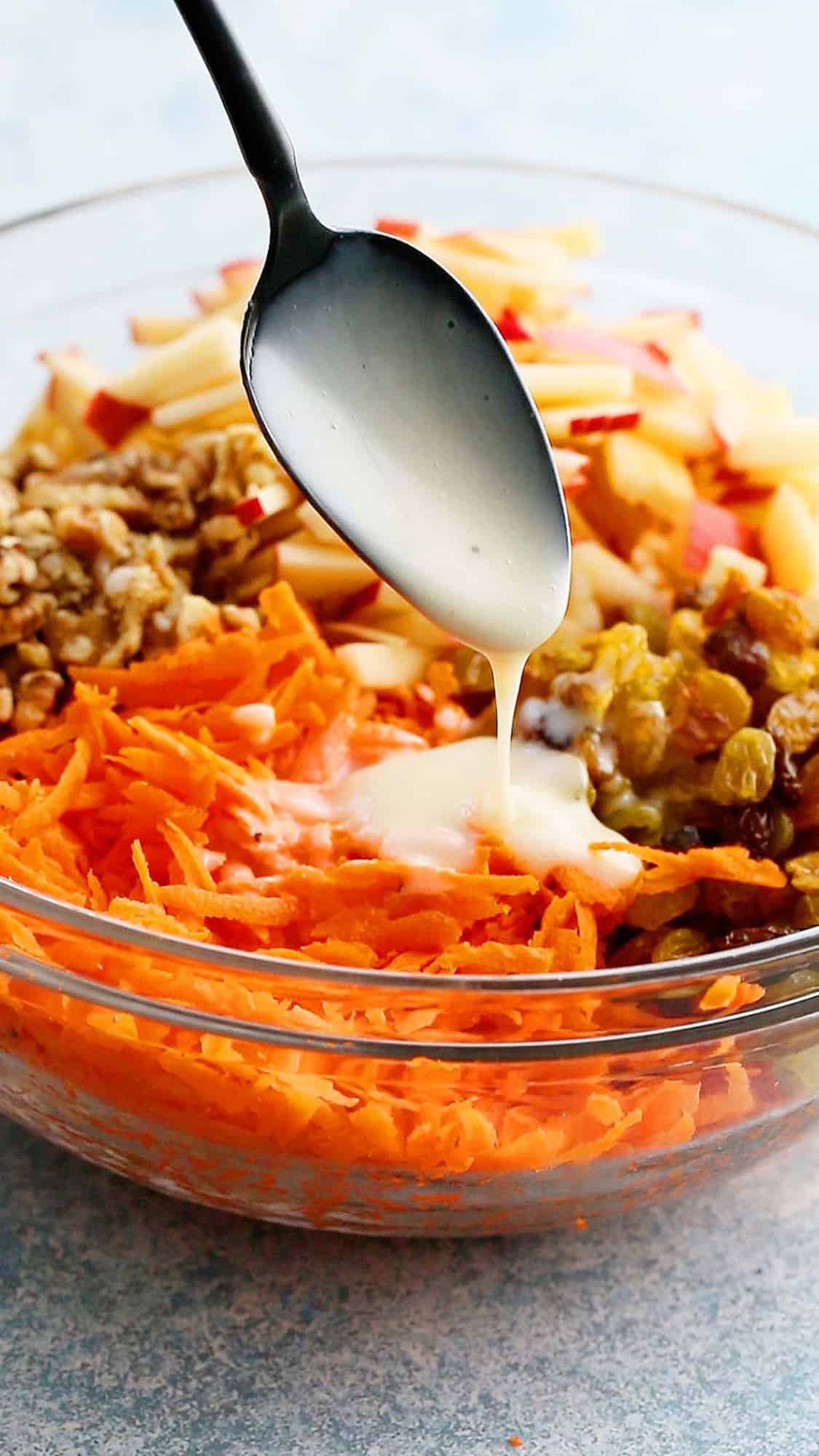 one black spoon drizzling white sauce into a glass bowl with carrot salad ingredients.