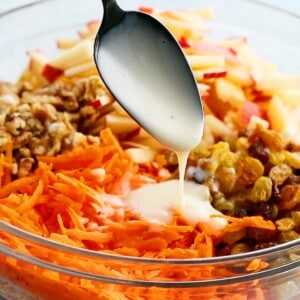 a black spoon drizzling white dressing over a glass bowl with carrot salad ingredients.