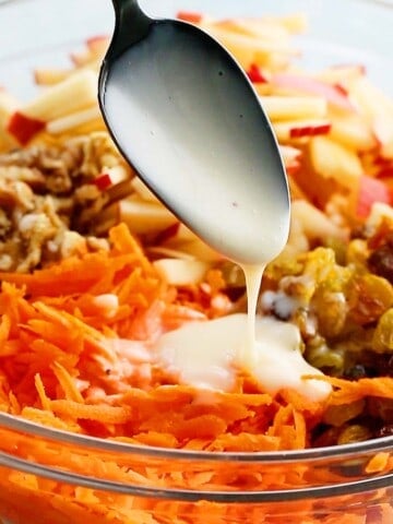 a black spoon drizzling white dressing over a glass bowl with carrot salad ingredients.