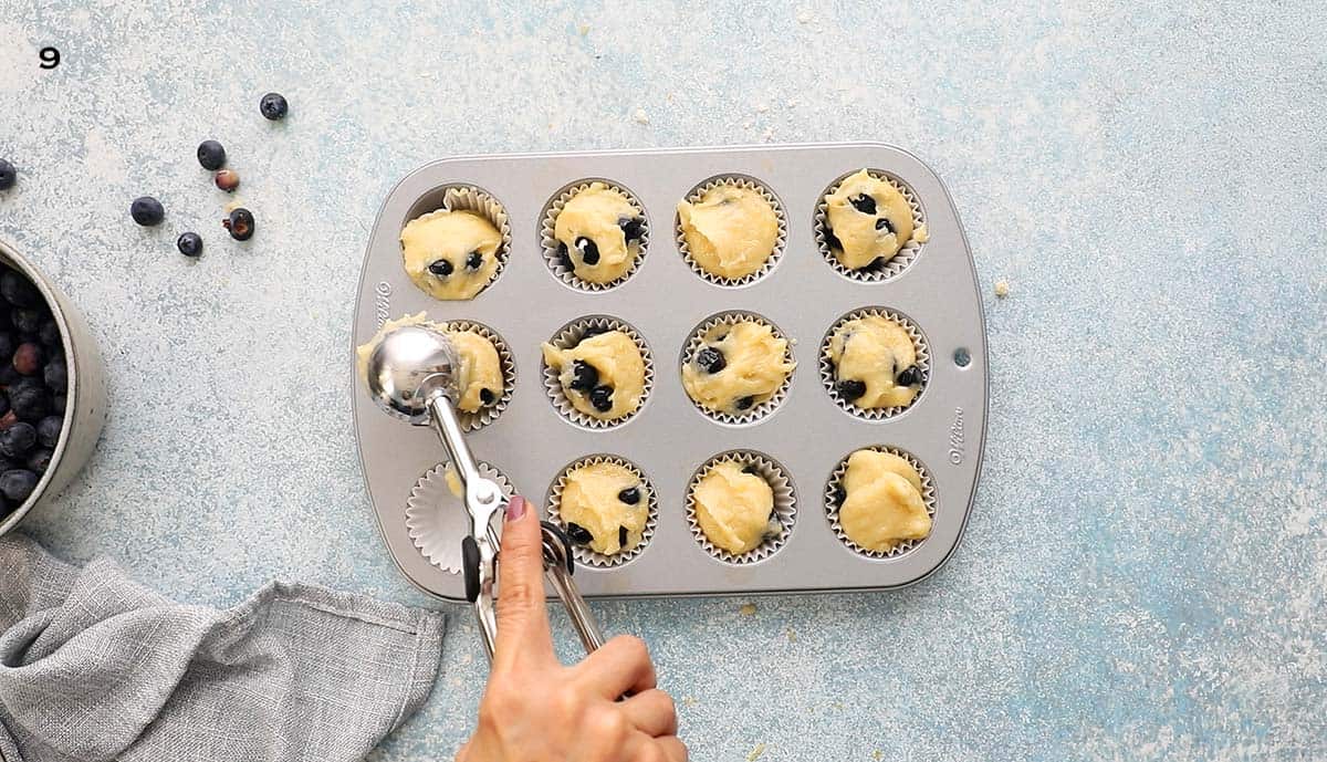 one hand adding batter into a 12-cup mini grey muffin pan.