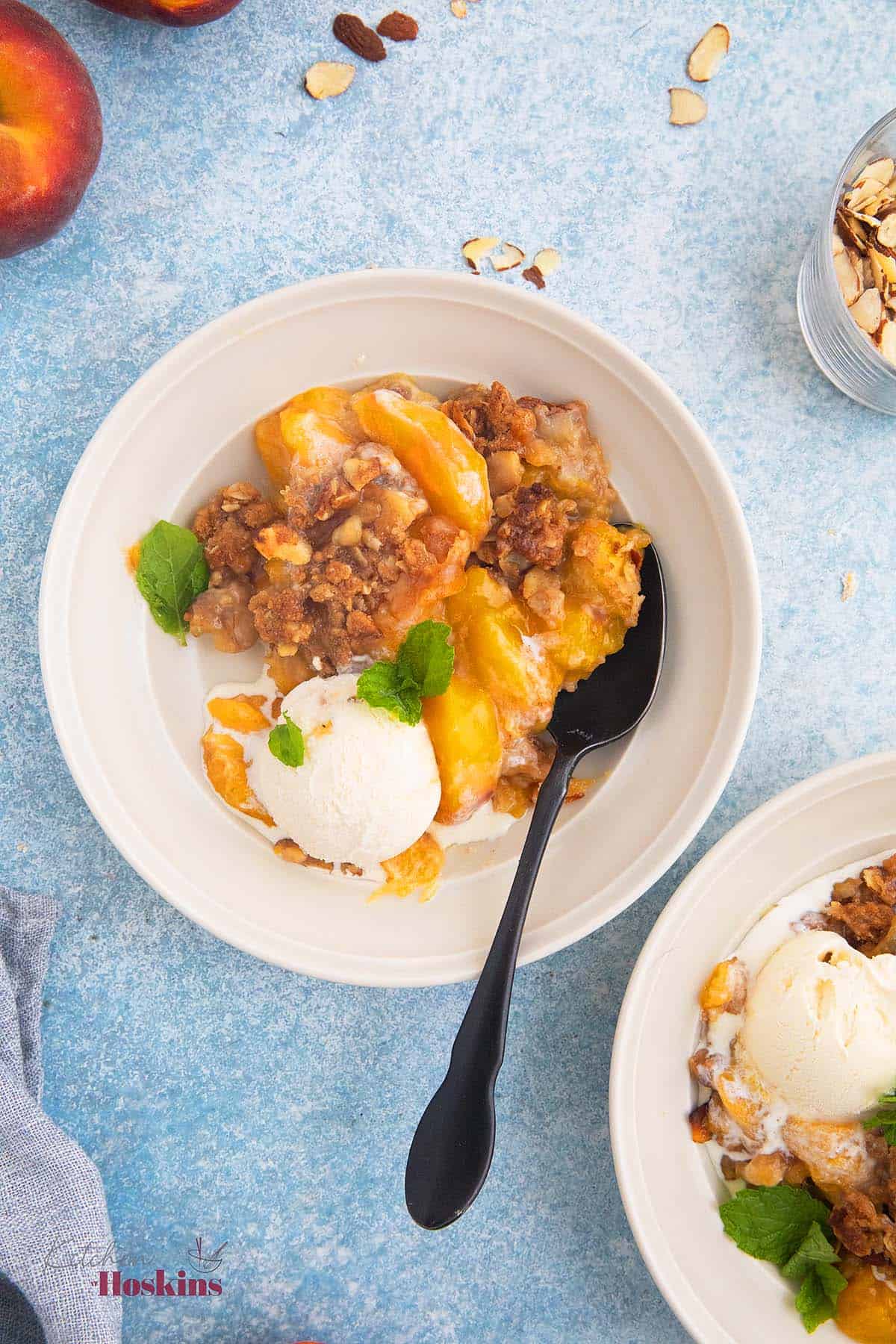 1 white bowl with peach crumble, one scoop of vanilla ice cream and a black spoon.