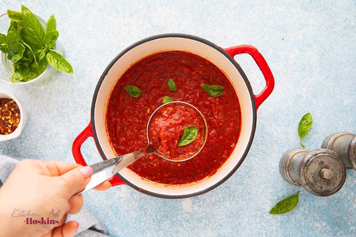 a hand lifting a ladle full of red sauce garnished with a basil leaf.