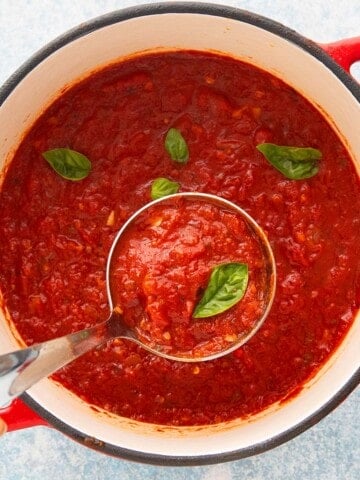 a ladle full of red sauce along with a basil leaf above a pot filled with the same.
