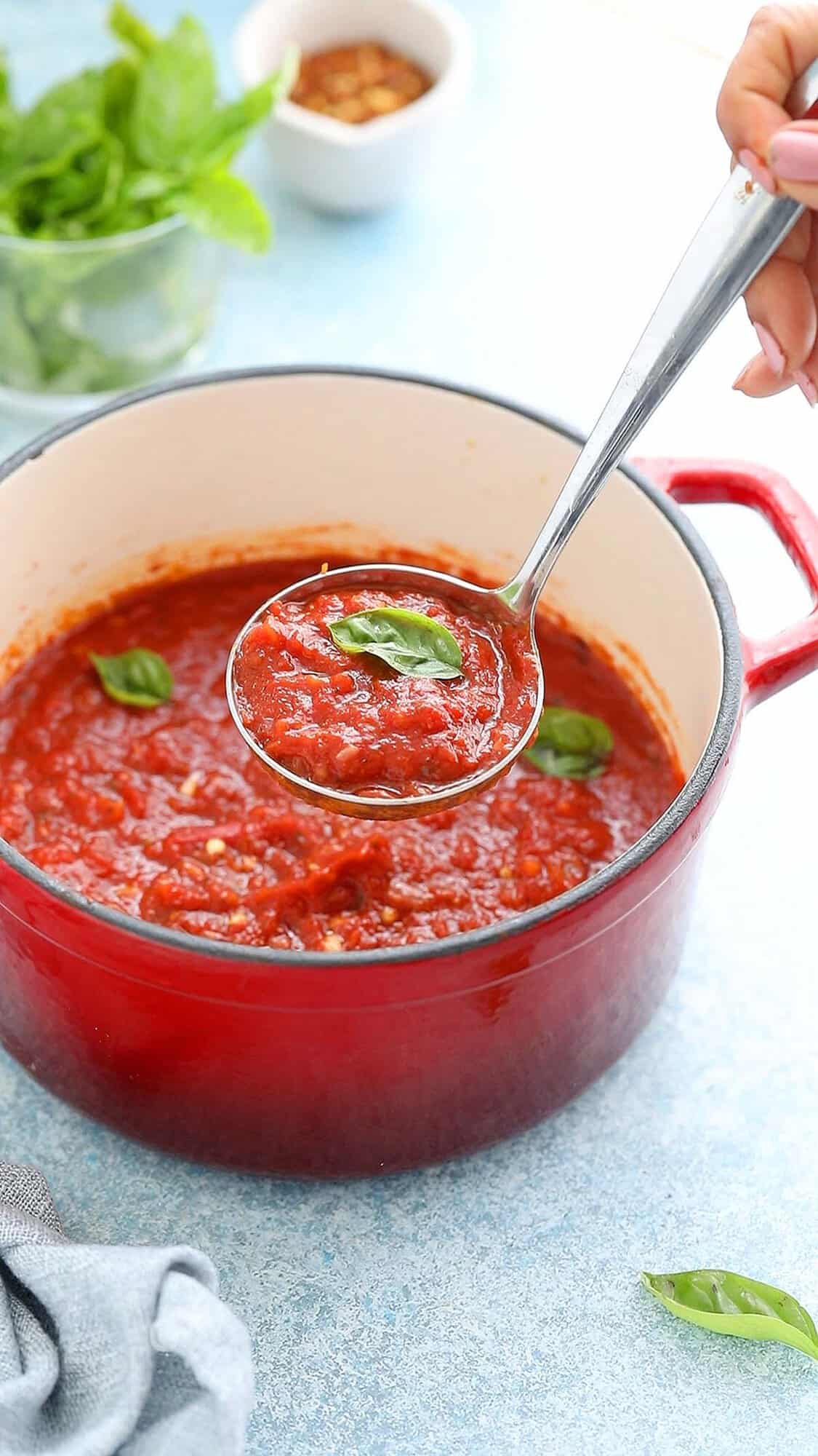 a hand lifting a ladle full of red sauce garnished with a basil leaf.