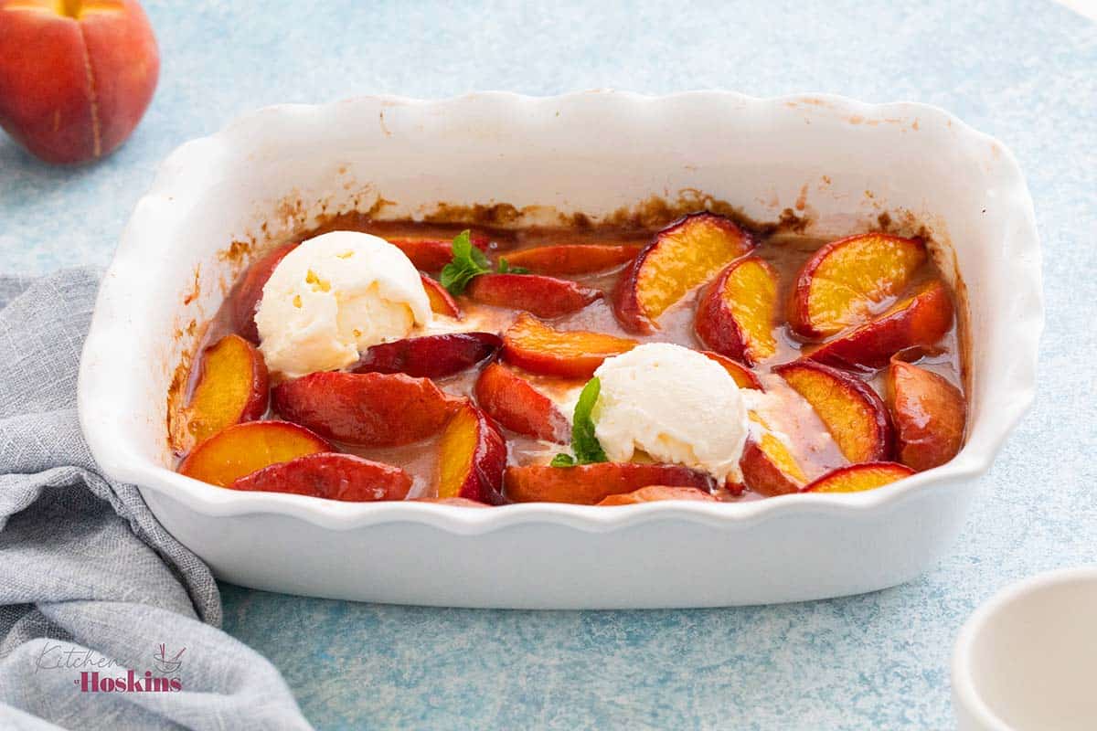 white fluted baking dish with baked peach wedges along with 2 scoops of vanilla ice cream.