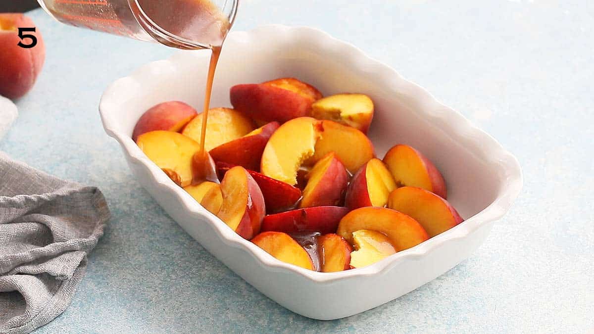 pouring brown liquid using a glass measuring cup into a white baking dish filled with peach wedges.