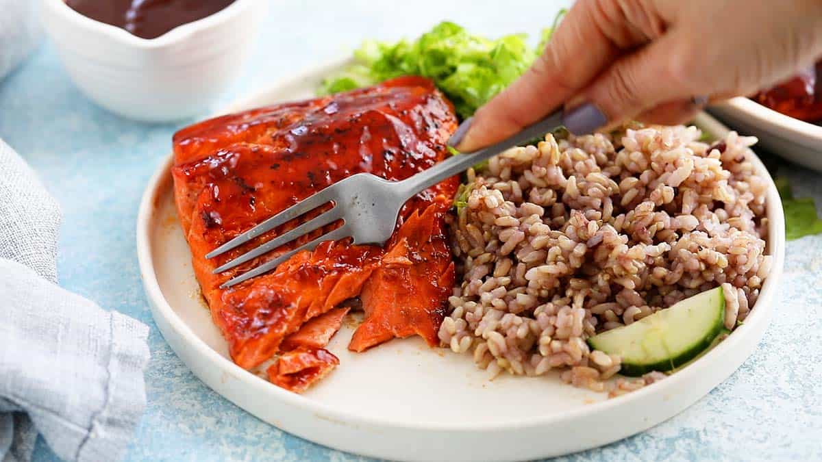 a hand using a fork to cut a piece of salmon from a white plate.