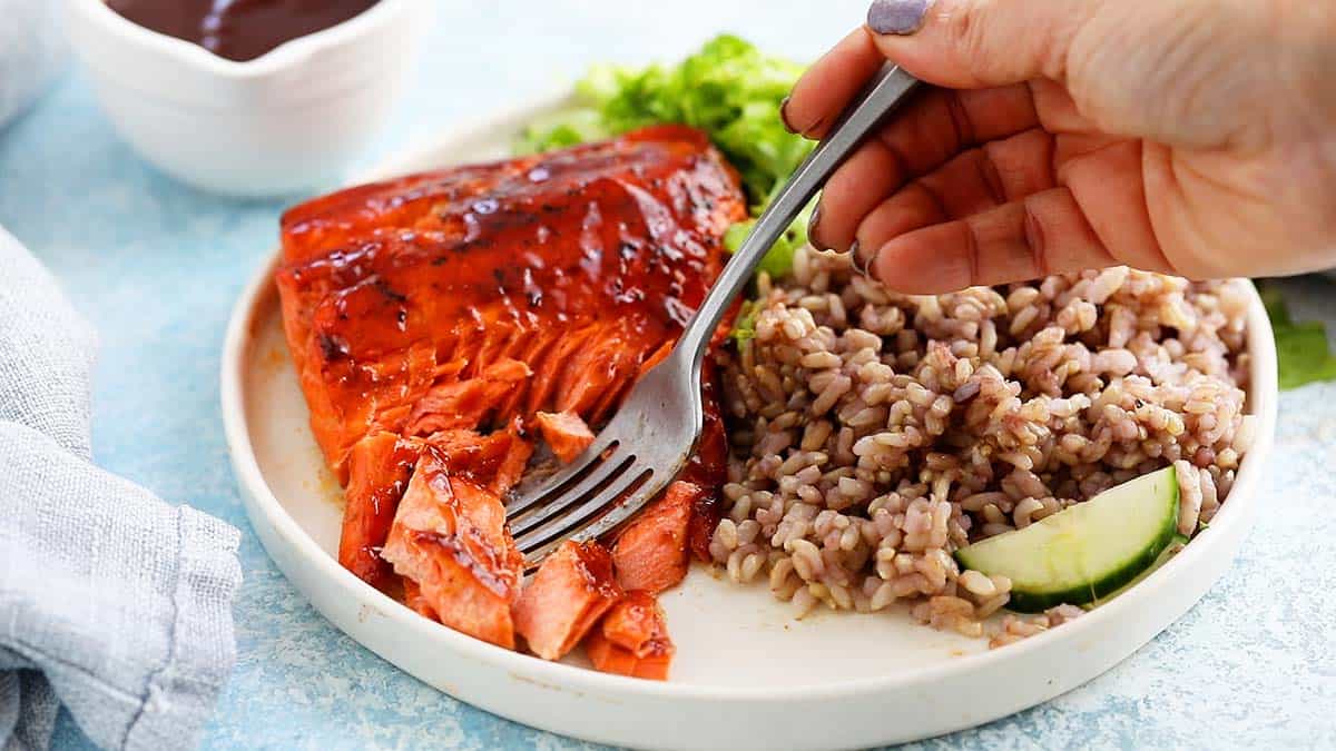 a hand using a fork to cut a piece of salmon from a white plate.