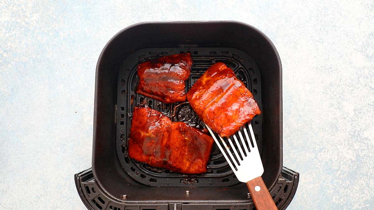 a hand lifting one cooked salmon fillet from an air fryer basket with a fish spatula.