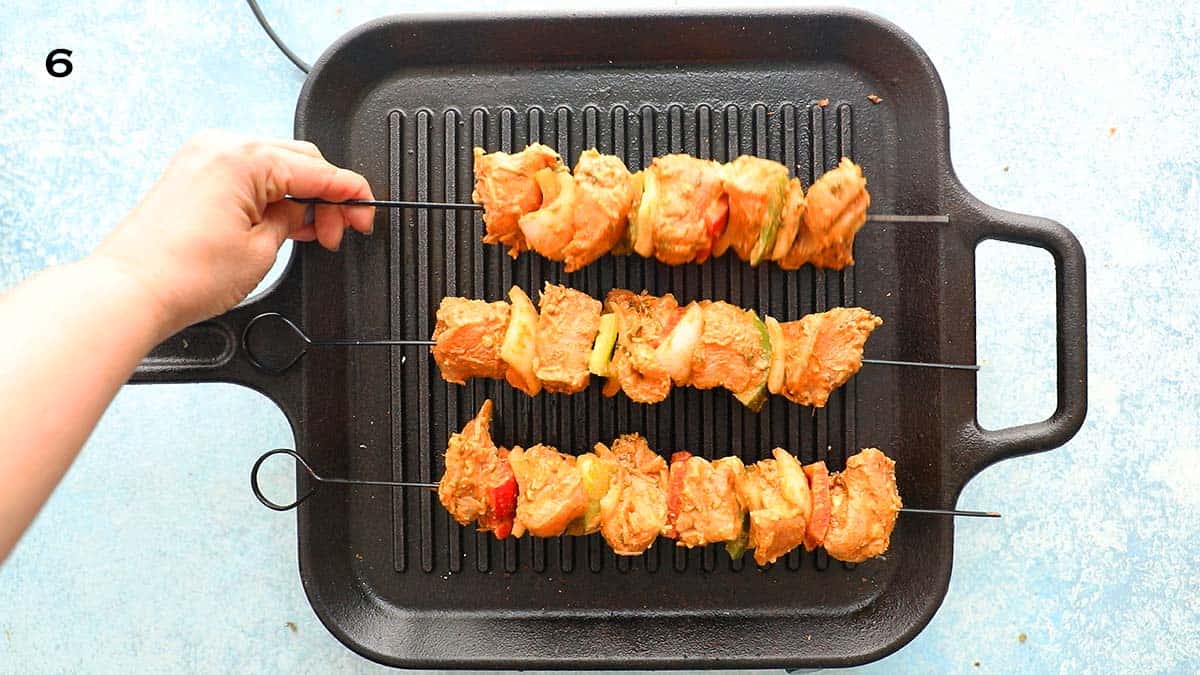 two hands cooking 3 skewered chicken kebobs on a black grill pan.