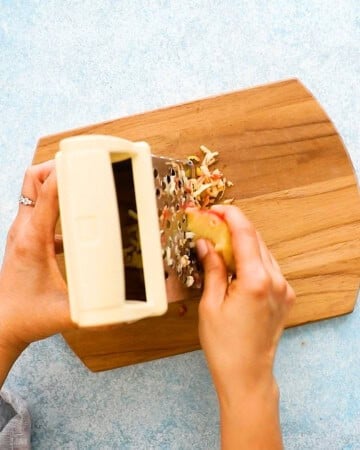 two hands grating one apple using a box grater.